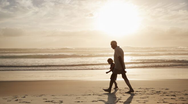 Silhouette Of Grandfather Walking Along Beach With Grandson